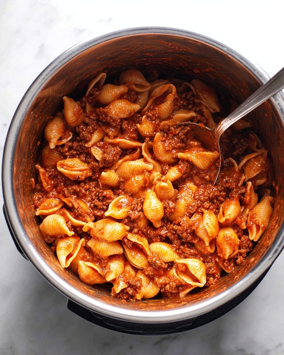 Inside a shiny metal pot, there is a mix of pasta shells and thick red sauce with chunks of cooked ground meat. The pasta is coated evenly with the sauce, giving it a deep orange-red color. The small shell pasta pieces show smooth textures and hold the sauce well. A metal spoon is partially dipped in the pot, mixing the pasta and meat sauce together. The pot sits on a white marbled surface. photo taken with an iphone --ar 4:5 --v 7