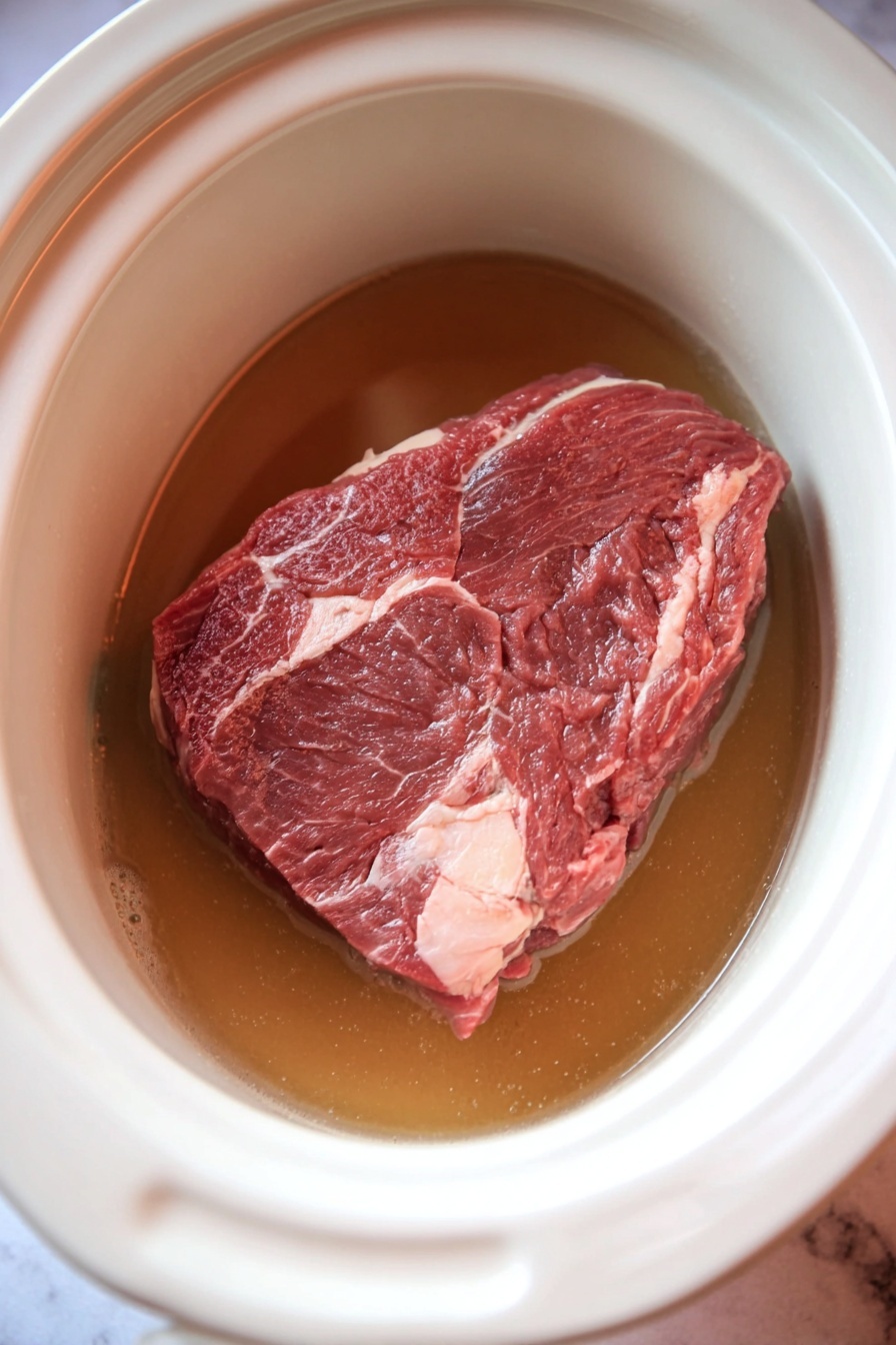 Slow Cooker Mississippi Roast, Mississippi Roast recipe, easy slow cooker dinner, tender beef roast, flavorful beef recipe - The image shows a white slow cooker filled with chunks of brown, cooked meat and several yellow-green whole peppers floating in a golden-brown oily sauce. The meat pieces vary in size and texture, some shredded and some solid, sitting below and partially above the surface of the liquid. The inside walls of the cooker have some sauce spills and splatters near the upper edge. The cooker is placed on a white marbled surface. Photo taken with an iphone --ar 2:3 --v 7