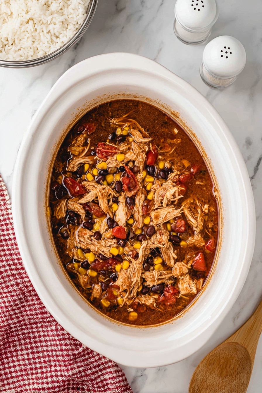 Slow Cooker Mexican Chicken, Mexican Chicken Recipes, Easy Mexican Chicken, Crockpot Mexican Chicken, Mexican Chicken Dinner Ideas - A white round plate is filled with a layer of fluffy white rice, topped in the center with shredded cooked chicken mixed with black beans, yellow corn, and small pieces of red tomato, giving a mix of brown, black, yellow, and red colors. A black fork rests on the right side of the plate, with some rice on its prongs. The plate is placed on a white marbled surface, with a white bowl of shredded chicken mix partially visible in the top left, two white salt and pepper shakers in the top right, a red and white checkered cloth in the bottom left corner, and a small white bowl filled with red cherry tomatoes in the bottom right corner. Photo taken with an iphone --ar 2:3 --v 7