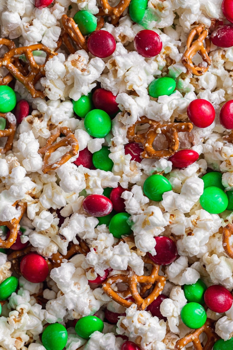 Christmas Crunch Popcorn, holiday popcorn snack, festive popcorn recipe, Christmas party snacks, easy holiday treats - This image shows a close-up of a holiday snack mix with three main layers closely mixed together. The first layer is white popped popcorn, fluffy and uneven in texture. Scattered among the popcorn are small twisted pretzels in a light brown color. The third layer has smooth, round candy pieces in bright red and green colors, spread evenly on top. The snack sits on a white marbled surface. photo taken with an iphone --ar 2:3 --v 7