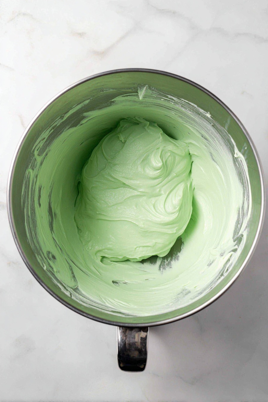 Christmas Tree Meringue Cookies, festive holiday cookies, Christmas dessert ideas, easy meringue cookies, holiday baking recipes - A shiny metal mixing bowl filled with smooth, thick light green frosting that has a creamy texture. The frosting sits in a rounded mound in the center of the bowl, with some streaks and swirls visible on its surface. The inside sides of the bowl have remnants of the frosting spread thinly, showing the mixing action. The bowl is placed on a white marbled surface, and part of the small metal handle is visible near the bottom right of the image. photo taken with an iphone --ar 2:3 --v 7