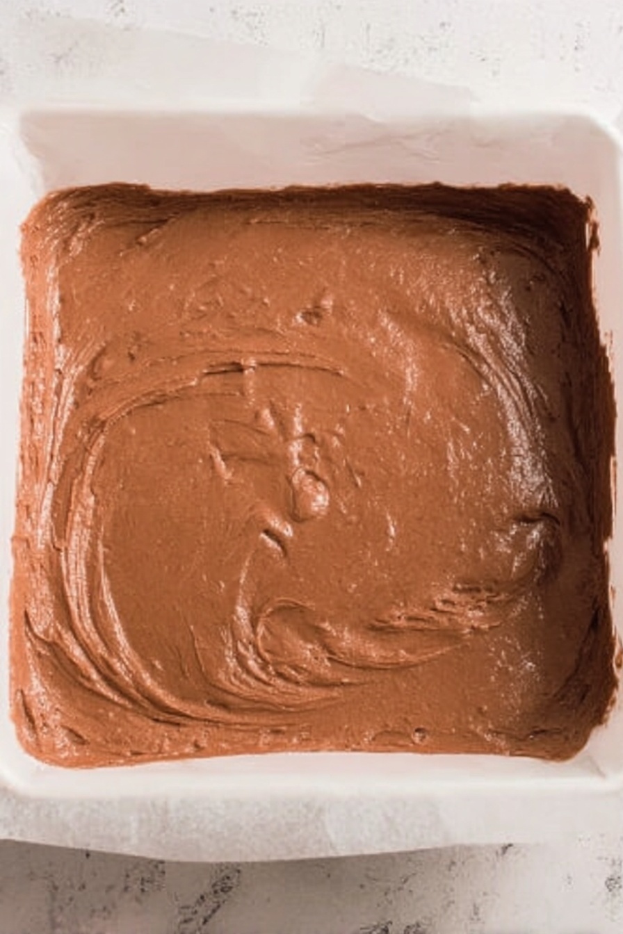 No-Bake Whipped Topping Chocolate Candy, chocolate candy recipe, easy no-bake desserts, quick chocolate treats, whipped topping chocolates - A white plate piled high with small square chocolate treats that have a smooth, glossy texture. One piece is on top with a bite taken out, showing a lighter brown, fluffy inside layer contrasting with the darker outer chocolate coating. Around the plate, more of these chocolate squares lay scattered on a white marbled surface, with a soft blue and white striped cloth partially under the plate. In the background, a white bowl holds several foil-wrapped chocolates, blurred softly to keep focus on the pile. photo taken with an iphone --ar 2:3 --v 7